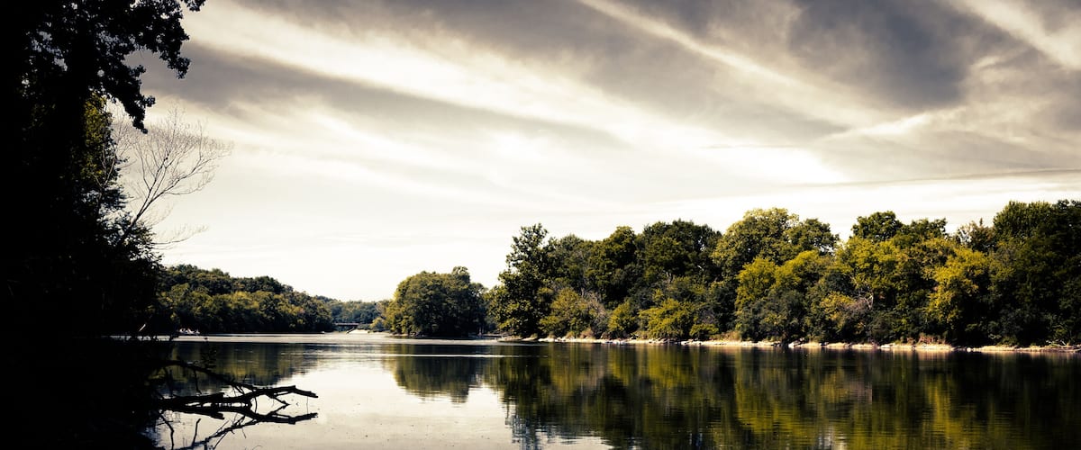 A calm lake with reflections of trees and clouds, surrounded by a forested area.