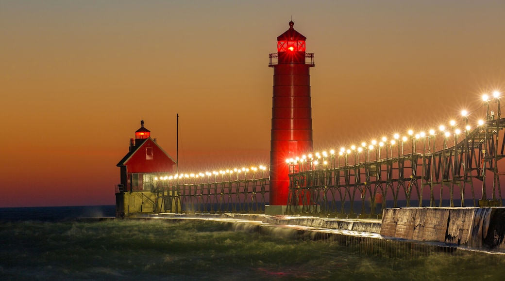 A red lighthouse with a lighted walkway leading to it, set against a sunset sky.