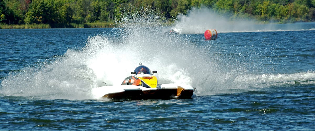 A speedboat creating a large splash on a lake with a forested shoreline in the background.