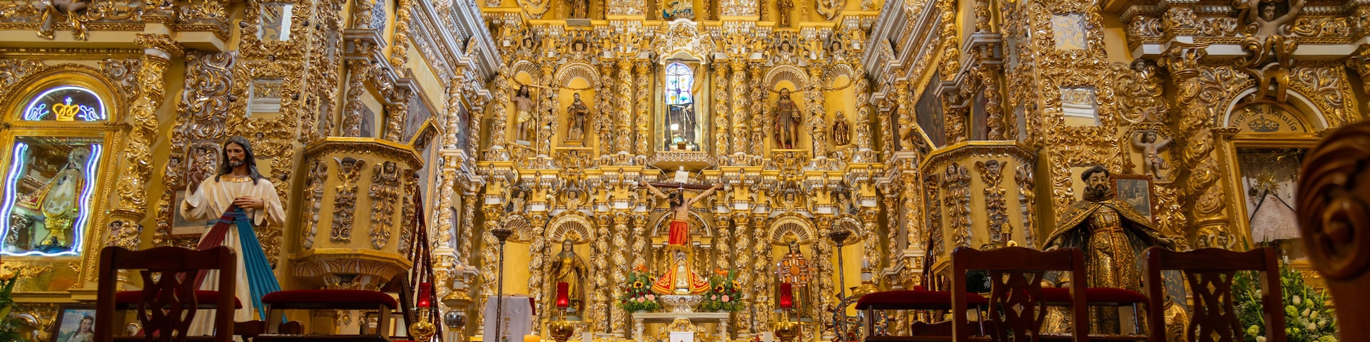 Interior de una iglesia ornamentada con decoraciones doradas, estatuas y sillas rojas.