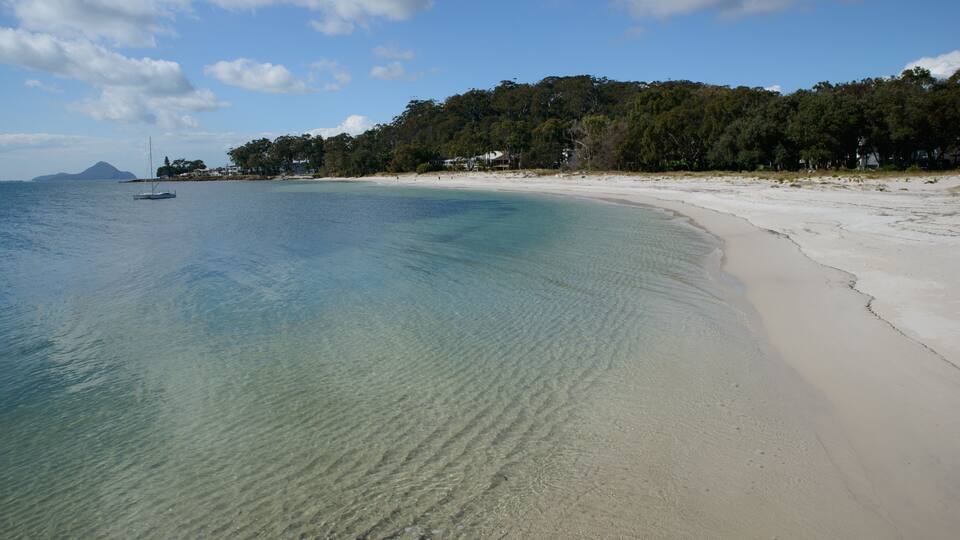 A beach with clear turquoise water, white sand, and a forested hill in the background.