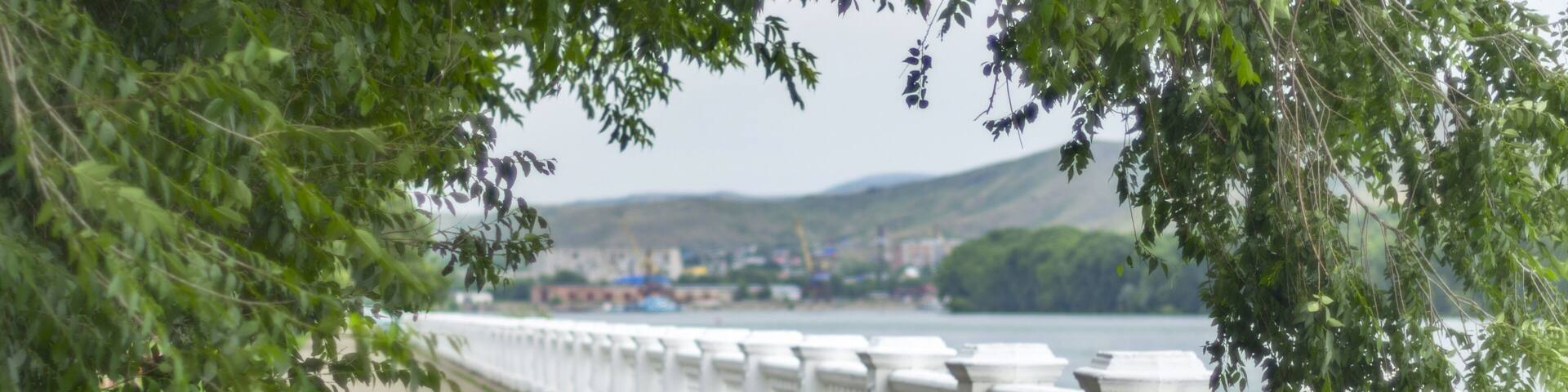Irtysh River Embankment Seen Through the Tree Crown Arch with Blurred Dockside Cranes, Apartment Blocks, Houses, and Mountains on a Summer Day, Ust-Kamenogorsk, Kazakhstan