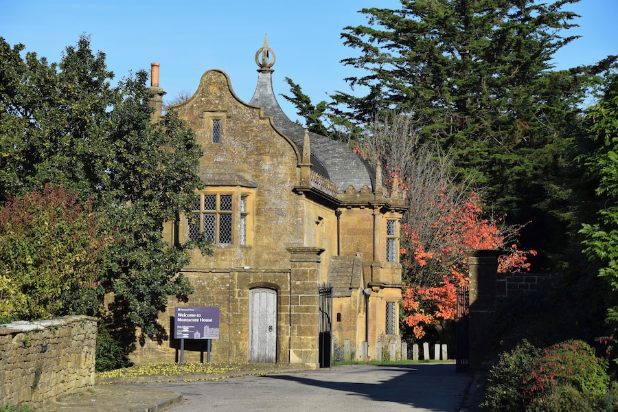 Un edificio de piedra histórico con un estilo arquitectónico tradicional, rodeado de árboles y un cartel de bienvenida a Montacute House.