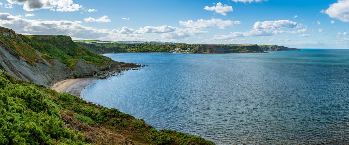 A coastal landscape with cliffs, a beach, and a clear blue sea under a partly cloudy sky.