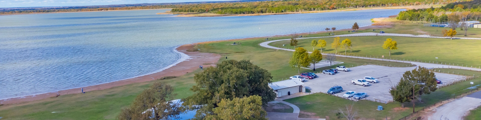 Un parque junto al lago con zona de estacionamiento, mesas de picnic y un pabellón.