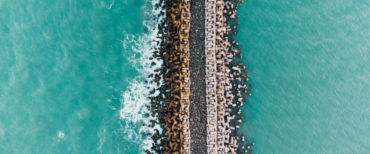 An aerial view of a coastal area with a long, narrow breakwater extending into the turquoise sea, surrounded by small rocks and pebbles.