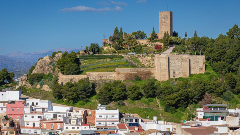 A historic city with a prominent tower and fortified walls on a hill, surrounded by residential buildings and lush greenery.