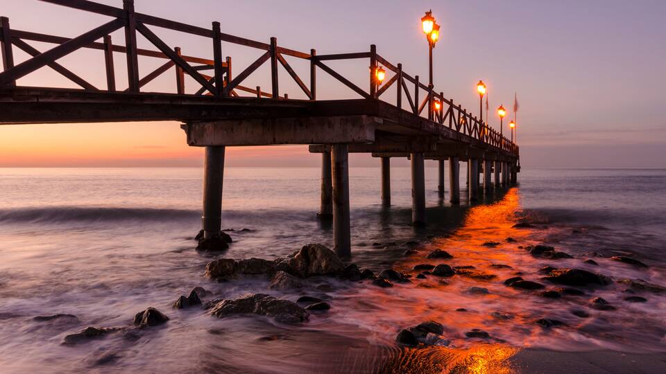 A wooden pier extending into the ocean at sunset with street lamps along its length.