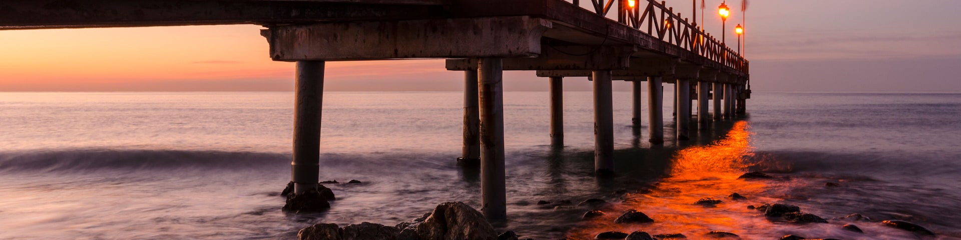 Un muelle de madera que se adentra en el océano al atardecer, con farolas a lo largo de su recorrido.