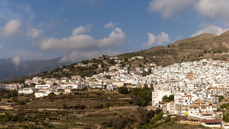 A densely packed hillside town with white buildings, surrounded by mountains and a clear sky.