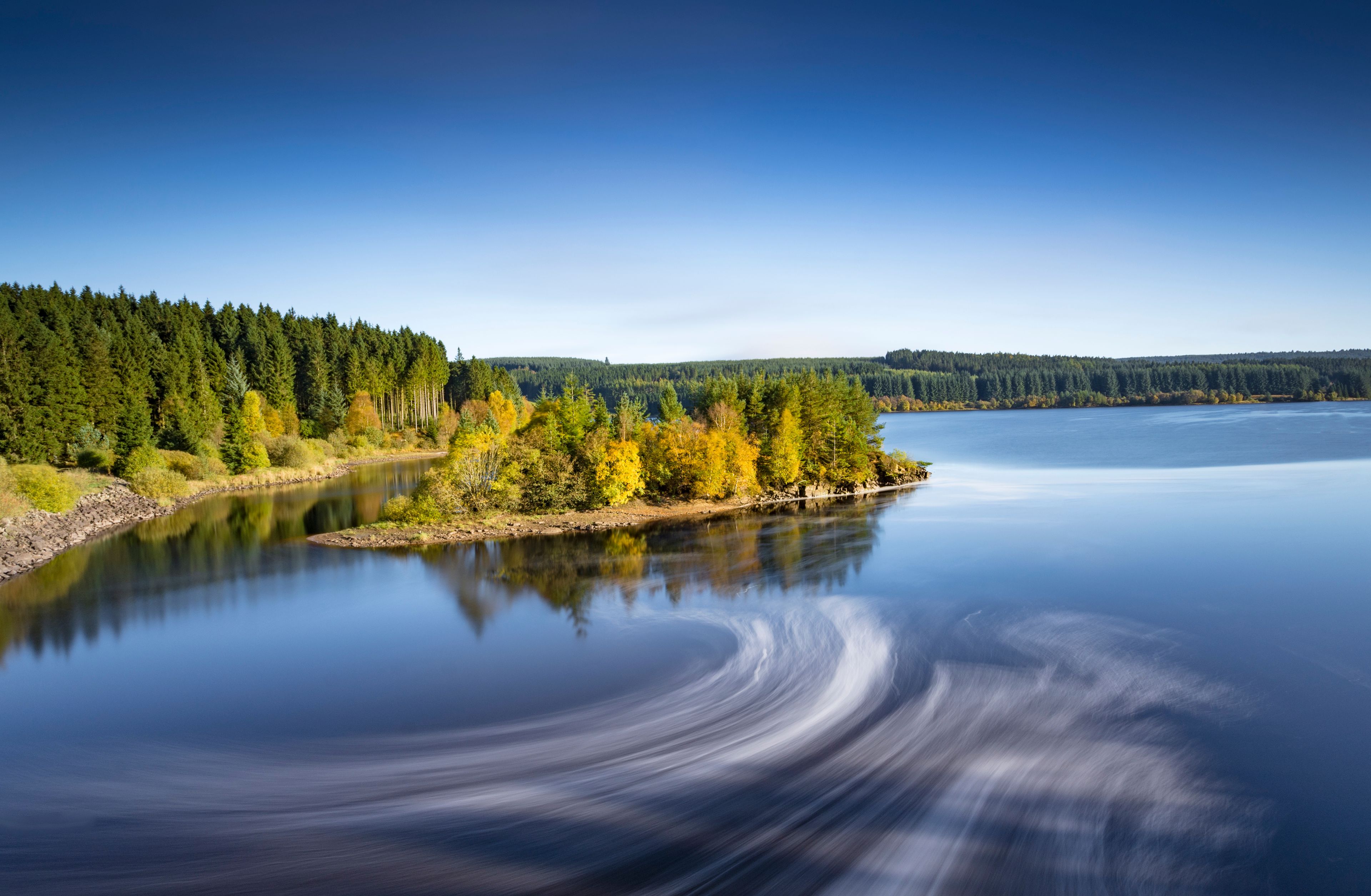 A calm lake with a reflection of the surrounding trees and a clear blue sky.