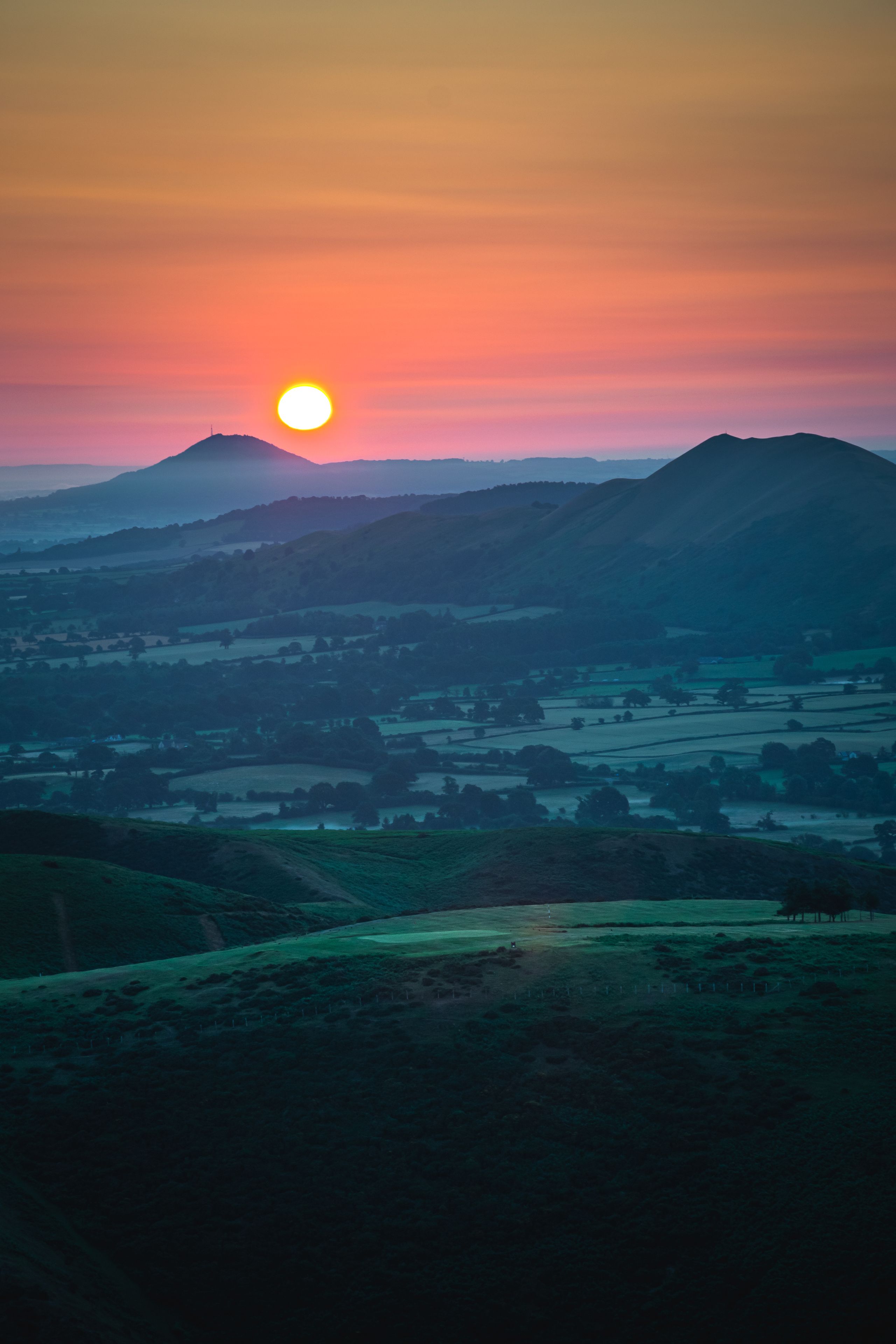 A sunset over a hilly landscape with a visible sun, distant mountains, and a valley with fields.