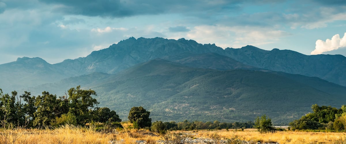 A mountainous landscape with a foreground of dry grass and scattered trees, and a range of mountains in the background under a partly cloudy sky.