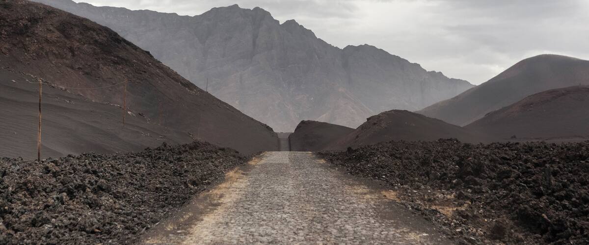 A gravel road flanked by barren hills and a mountain range in the distance.