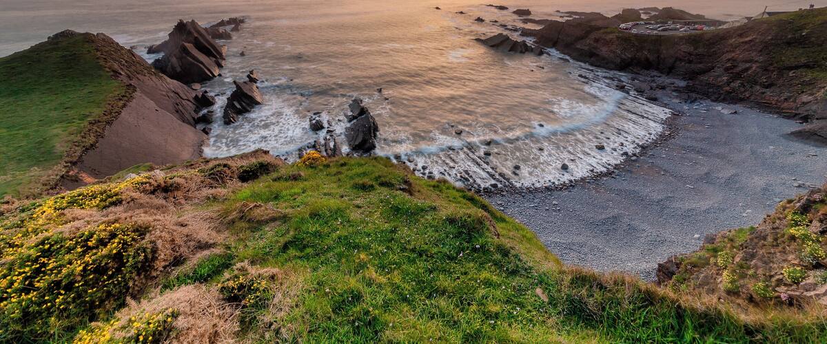 A coastal landscape with rocky cliffs, a pebbly beach, and a sunset over the ocean.