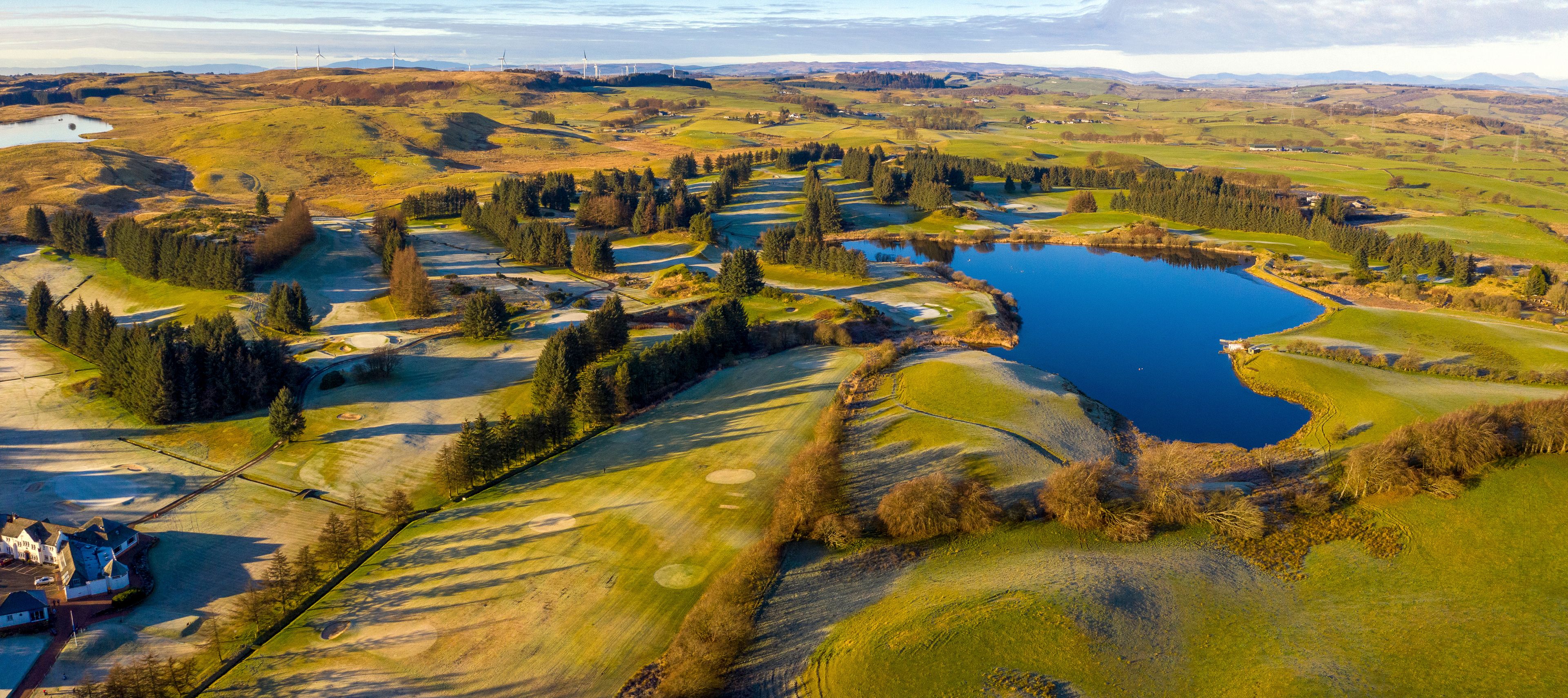 Un campo da golf con un lago, circondato da dolci colline e pale eoliche in lontananza.