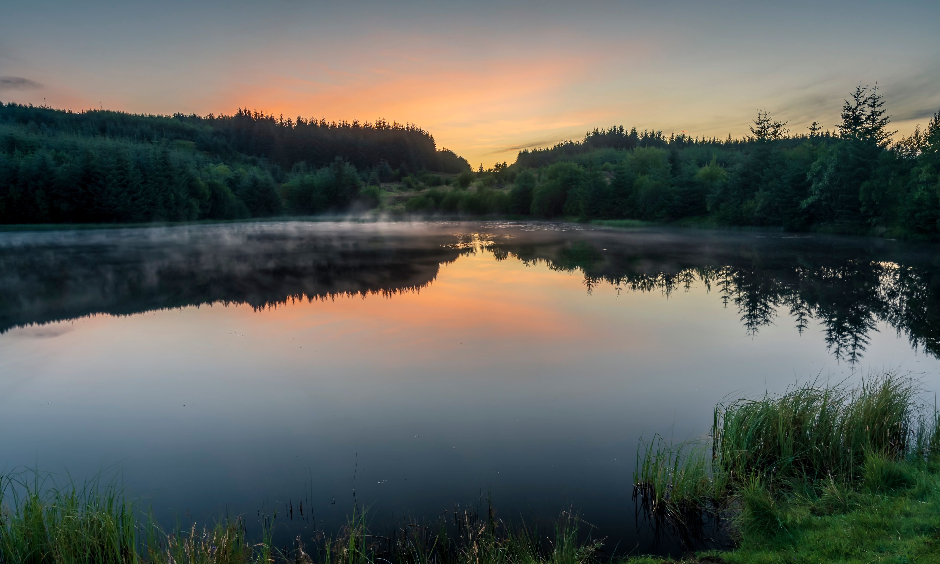 Een rustig meer dat bomen en een bebost helling weerspiegelt bij zonsopkomst of zonsondergang.