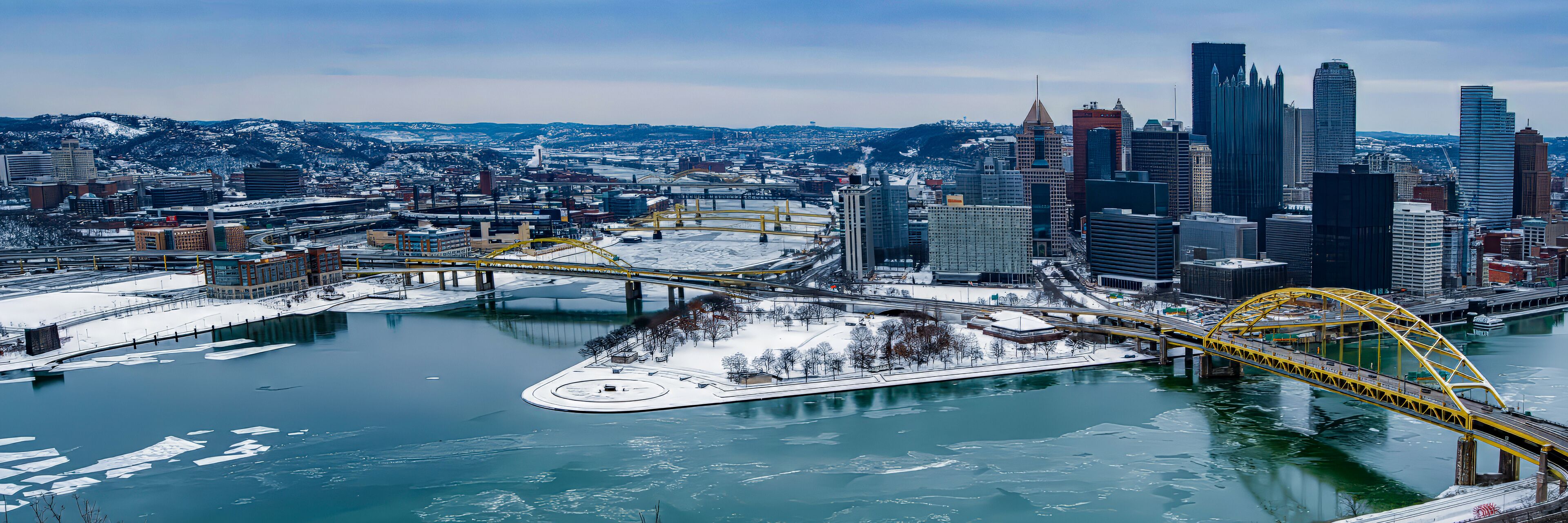 A cityscape with a prominent yellow bridge over a frozen river, surrounded by modern skyscrapers and snowy hills in the background.