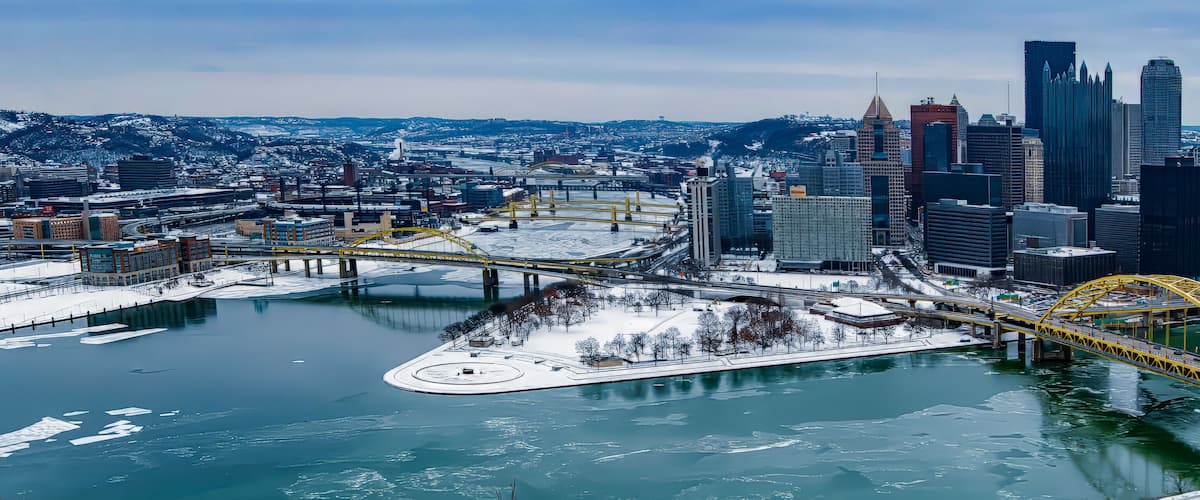 A cityscape with a prominent yellow bridge over a frozen river, surrounded by modern skyscrapers and snowy hills in the background.