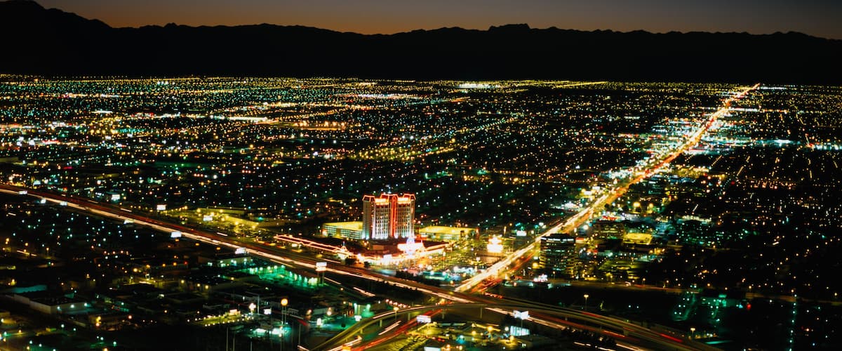 A cityscape at night with a prominent building with red lights, numerous roads, and a mountainous backdrop.