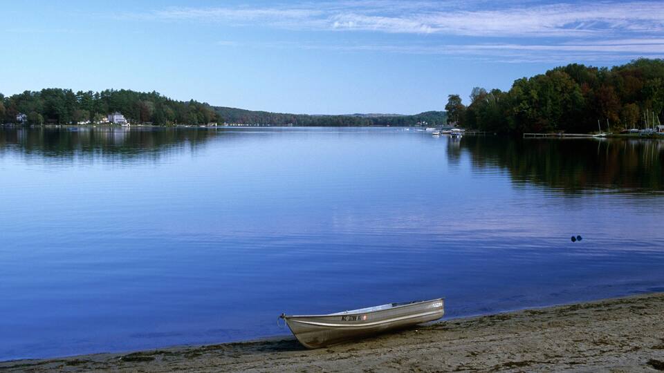 A calm lake with a boat on the shore and a forested hill in the background.