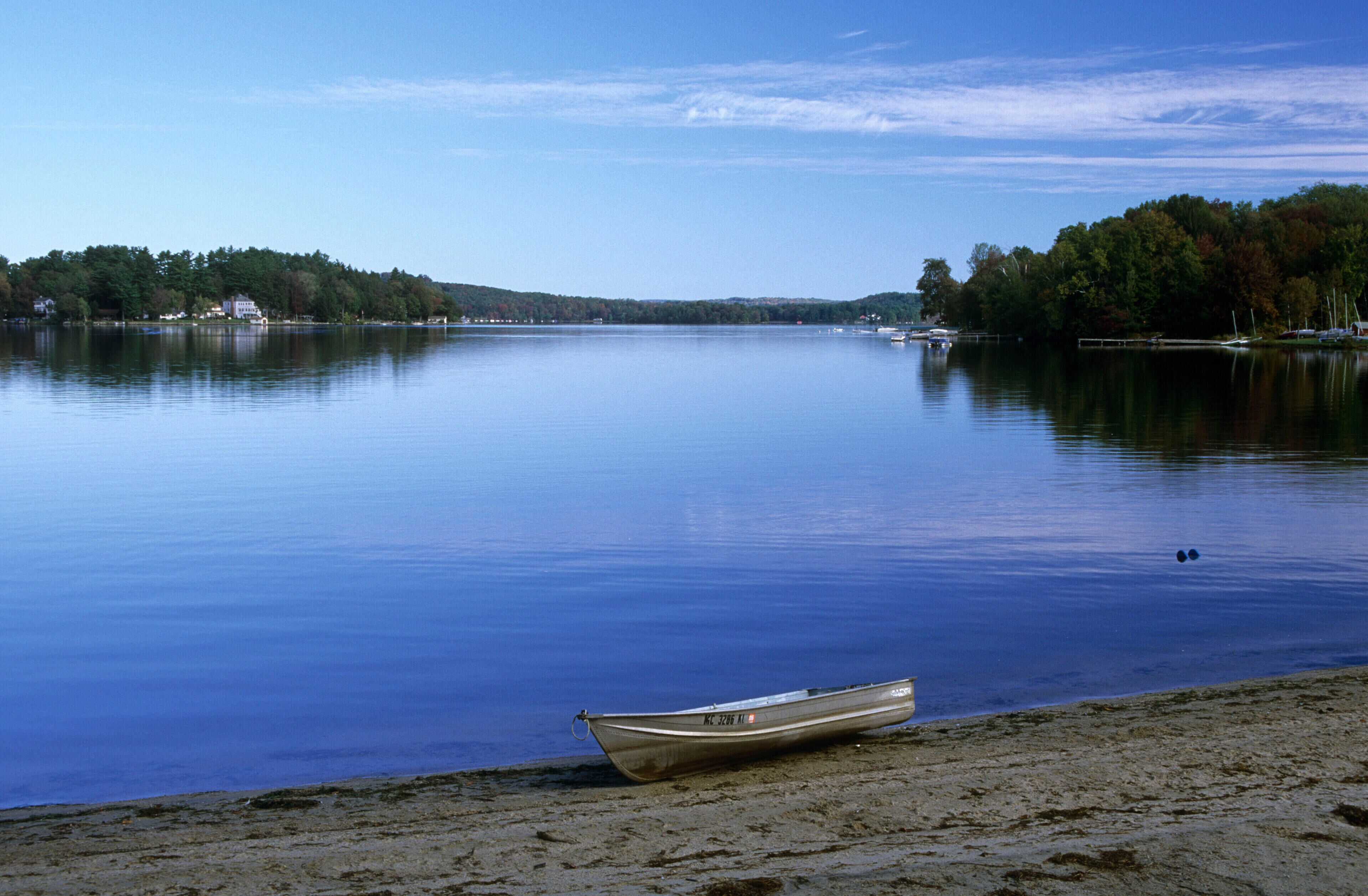 A calm lake with a boat on the shore and a forested hill in the background.