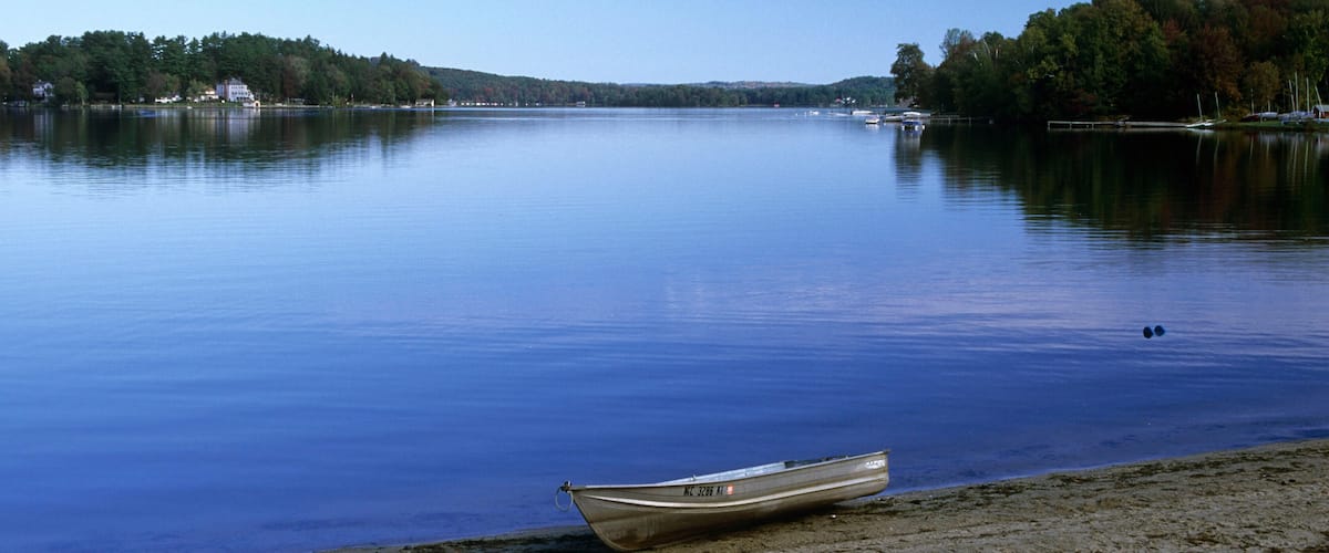 A calm lake with a boat on the shore and a forested hill in the background.