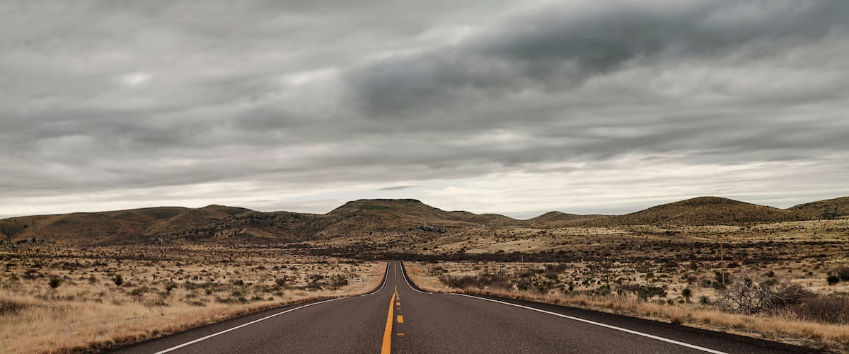 A road leading through a desert landscape with hills in the distance under a cloudy sky.