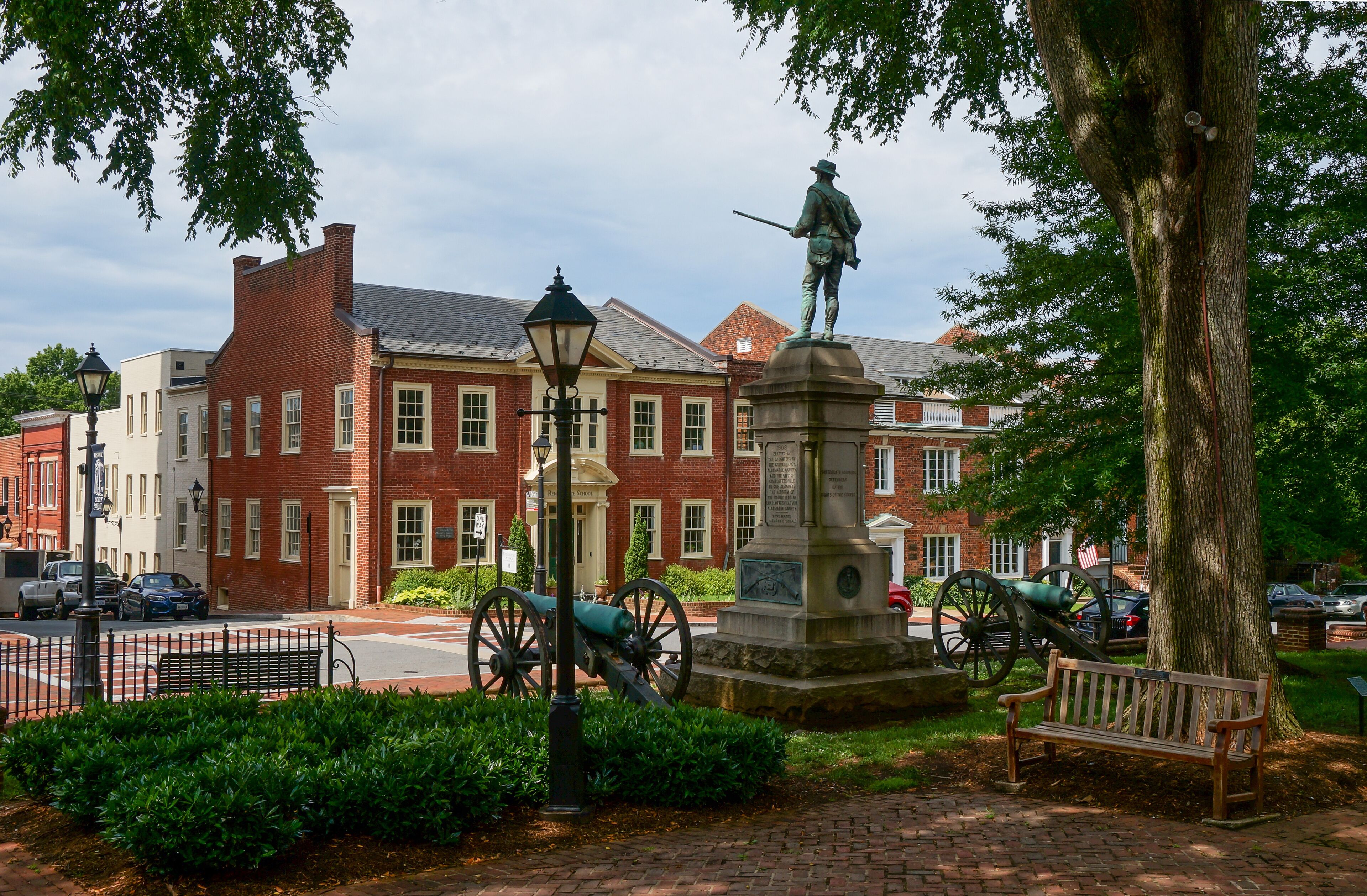 A statue of a historical figure, a cannon, and a bench in a park with brick buildings in the background.