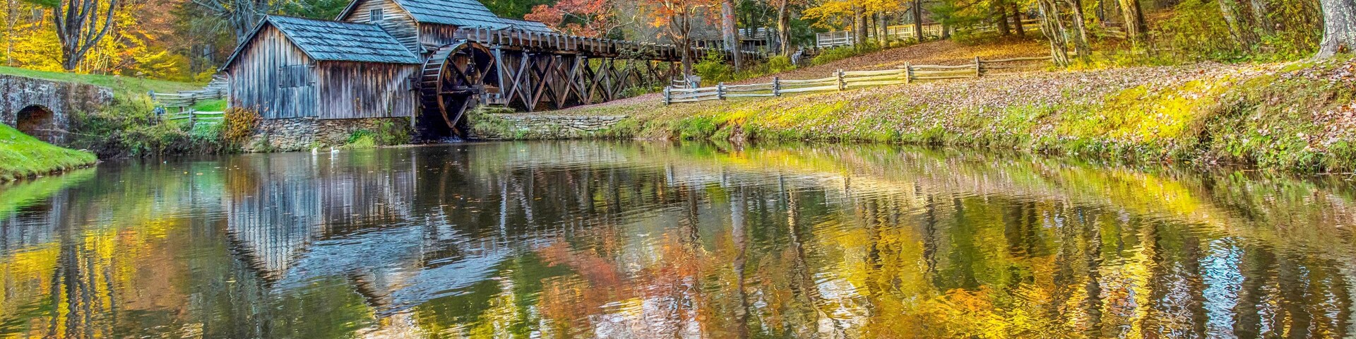 A wooden watermill with a stone bridge, surrounded by autumn-colored trees and a calm river.