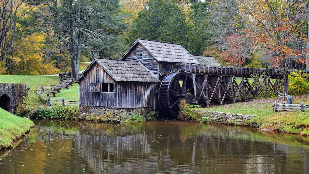 Un moulin en bois doté d’une roue à eau, entouré d’un feuillage automnal et bordé par une rivière paisible.