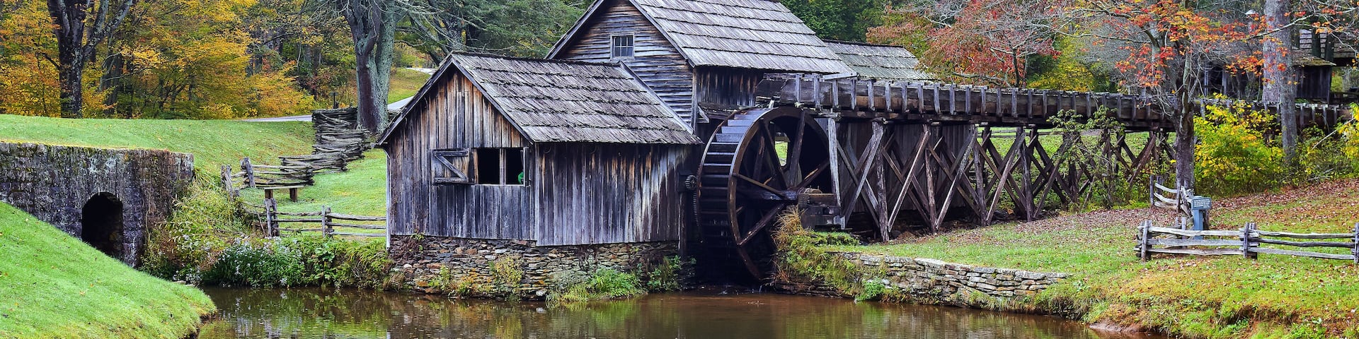 A wooden mill with a water wheel, surrounded by autumn foliage and a calm river.
