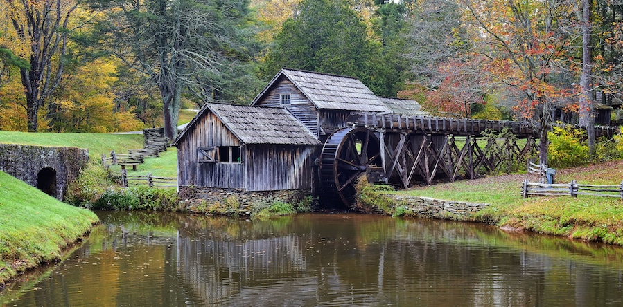Eine hölzerne Mühle mit Wasserrad, umgeben von Herbstlaub und einem ruhigen Fluss.