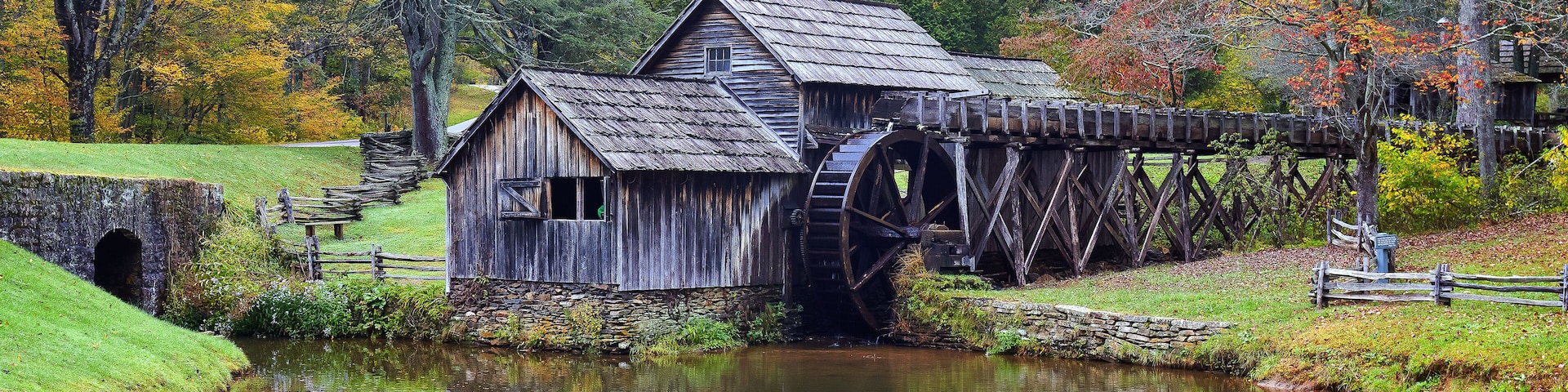Un moulin en bois doté d’une roue à eau, entouré d’un feuillage automnal et bordé par une rivière paisible.