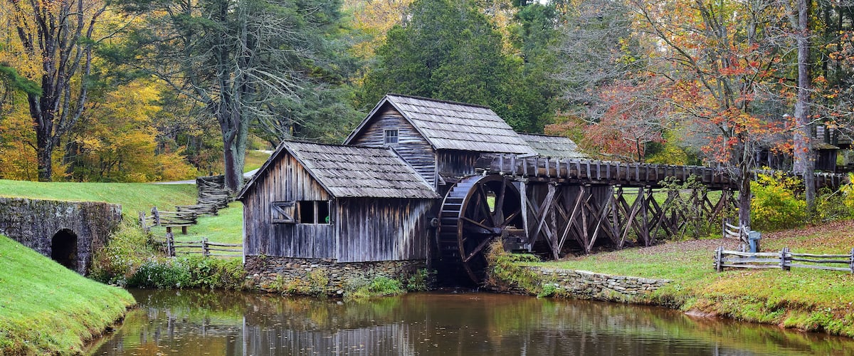 A wooden mill with a water wheel, surrounded by autumn foliage and a calm river.