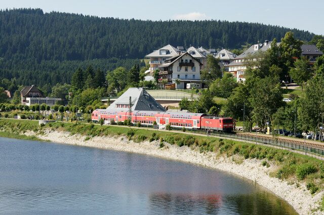 A lake with a red train on the tracks, surrounded by houses and trees.