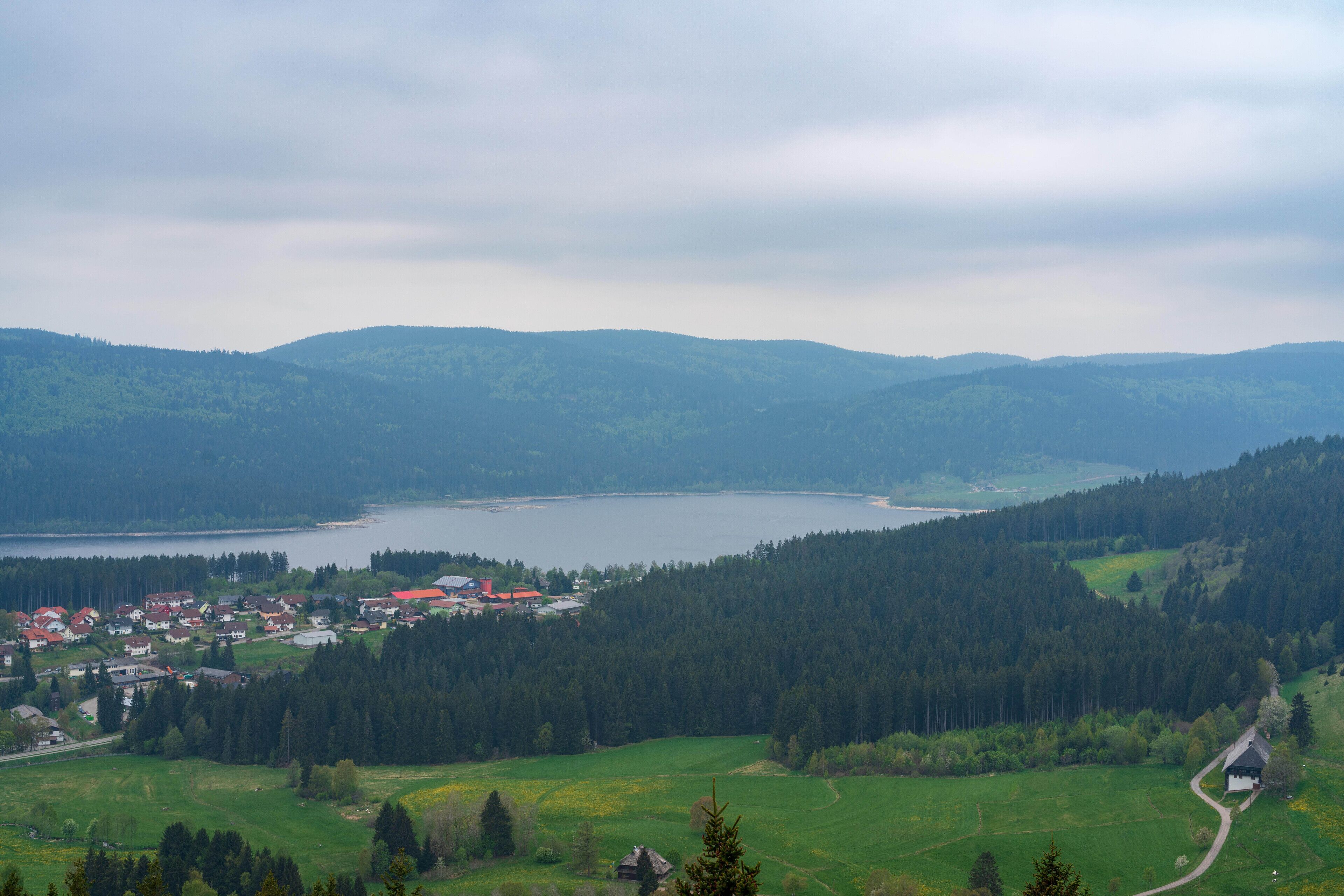 A lake surrounded by forested hills, a small village with red-roofed houses, and a winding road.