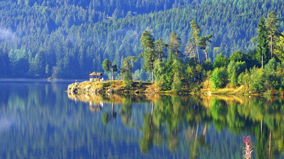 A calm lake with a small house on a rocky outcrop surrounded by dense forest.
