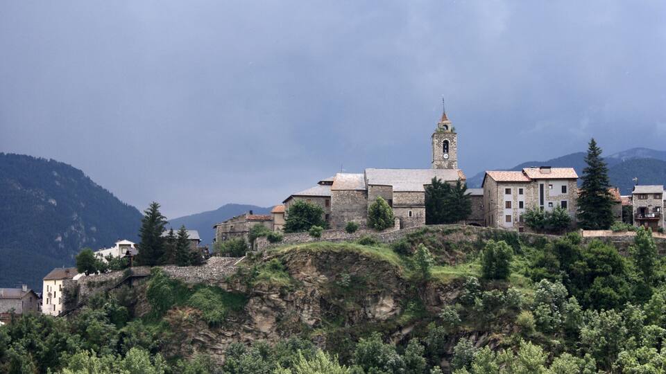 A historic town with a prominent church tower, surrounded by lush greenery and mountains in the background.