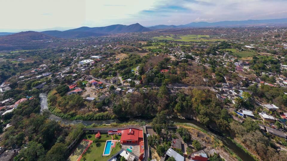 An aerial view of a town with a river, green hills, and a large swimming pool.