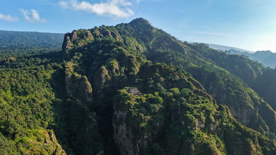 A mountainous landscape with dense green forests and a small ancient structure on a cliff.