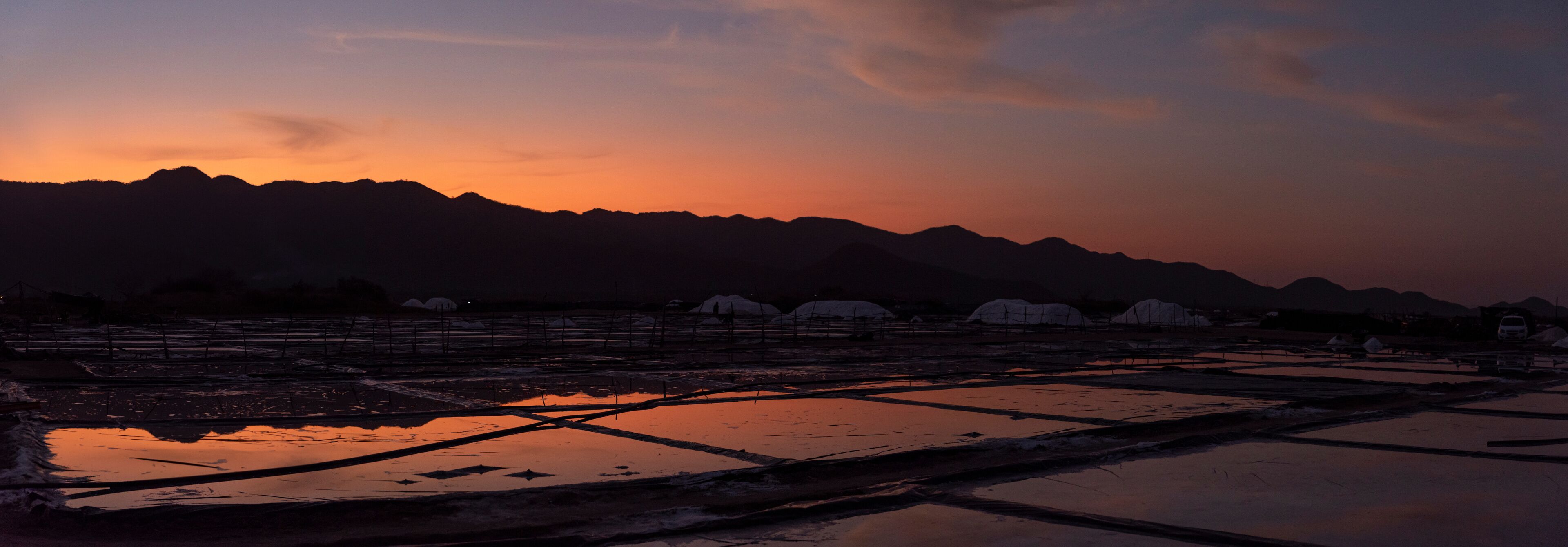 A sunset over a mountainous landscape with reflective water bodies and snow-covered structures.