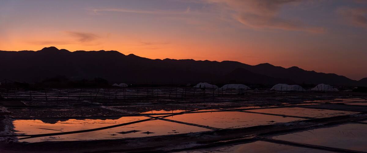 A sunset over a mountainous landscape with reflective water bodies and snow-covered structures.
