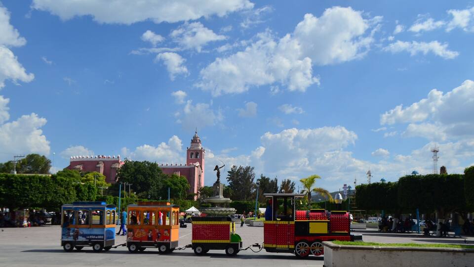 A colorful train in a park with a large building and a statue in the background.