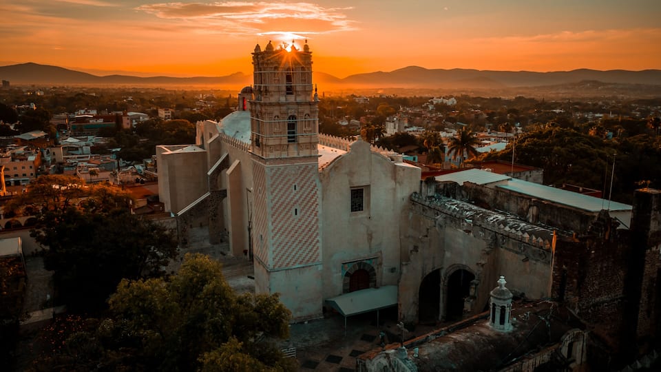A historic church with a bell tower, surrounded by urban buildings and palm trees, during sunset.