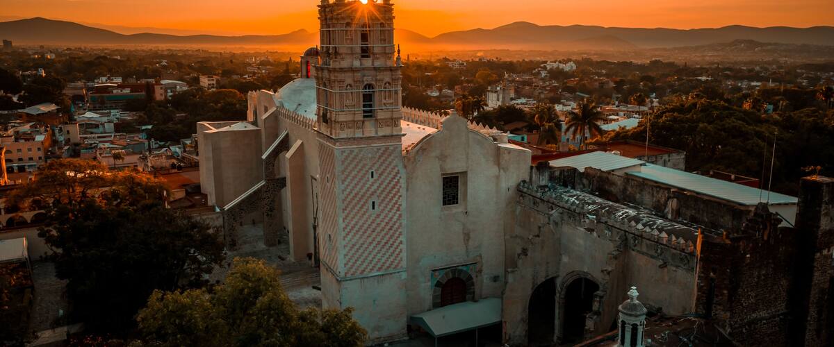 A historic church with a bell tower, surrounded by urban buildings and palm trees, during sunset.