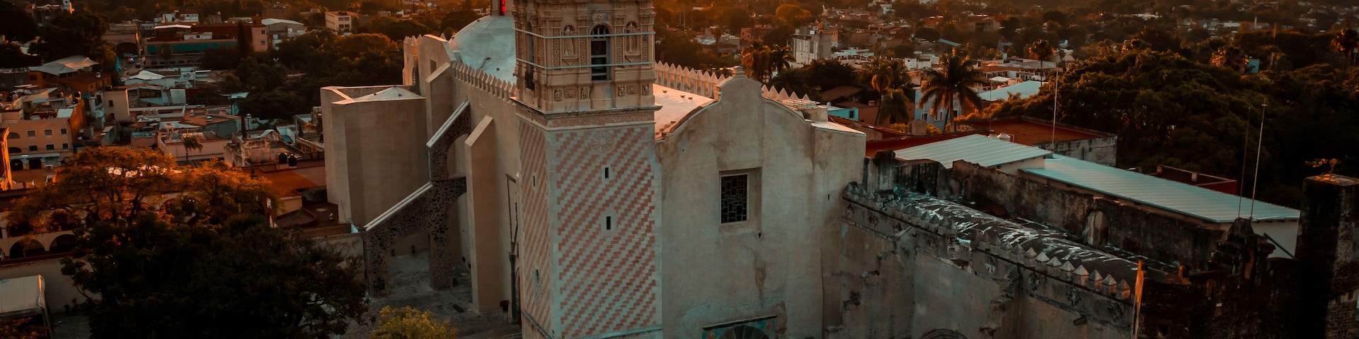A historic church with a bell tower, surrounded by urban buildings and palm trees, during sunset.