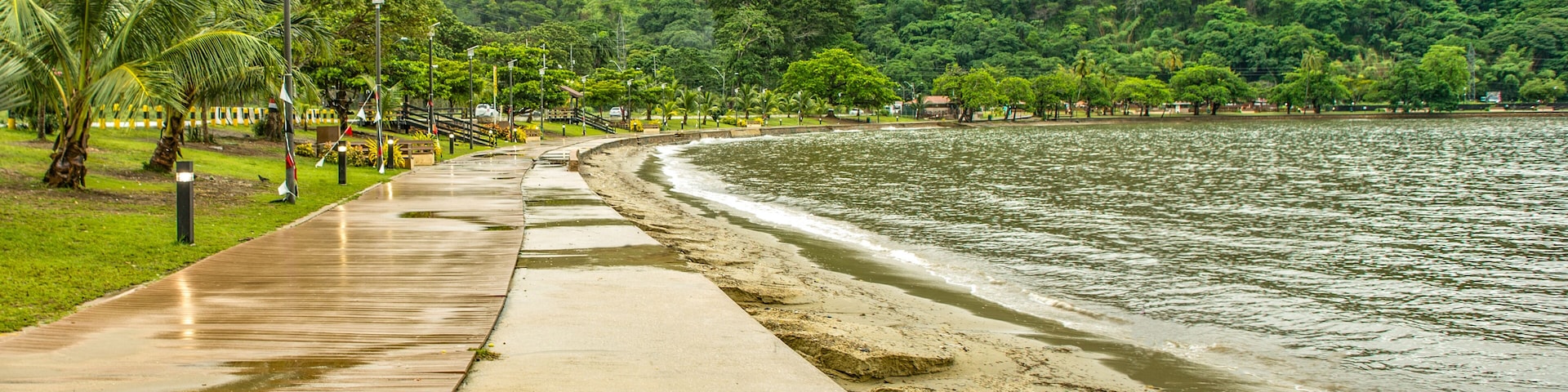 A coastal walkway with palm trees, a sandy beach, and a forested hill in the background.