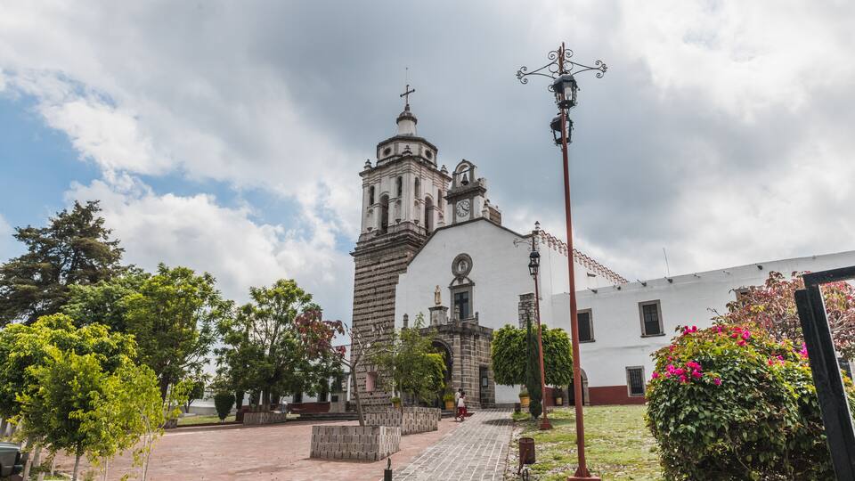 A historic church with a bell tower, surrounded by a paved courtyard with trees and a street lamp.