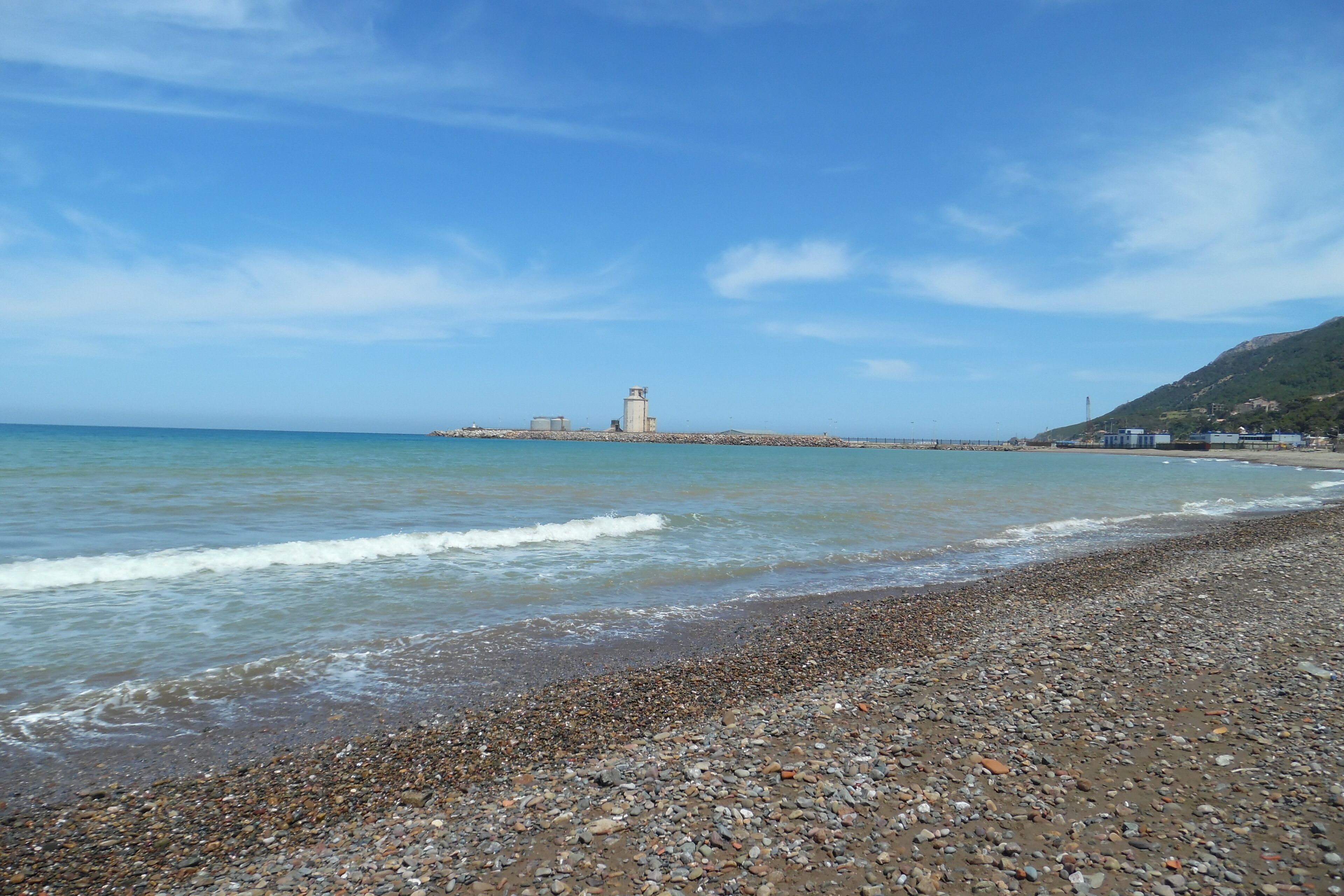 A pebbled beach with gentle waves, a distant lighthouse, and a hilly landscape.